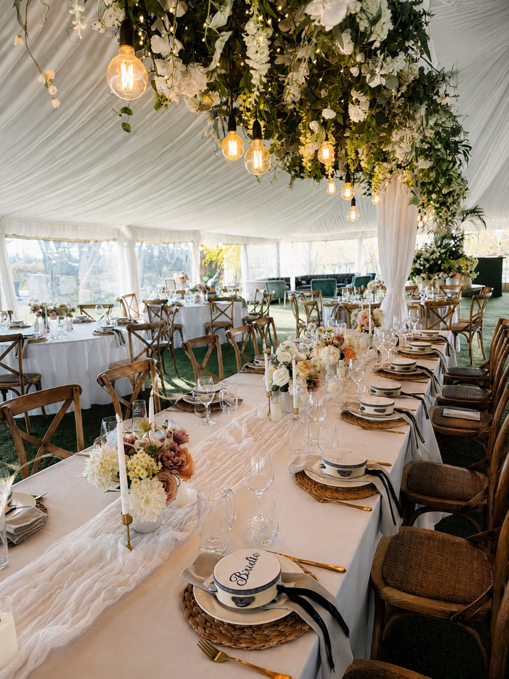 Wedding marquee with a long table, cross-back chairs, festoon bulbs and hanging greenery